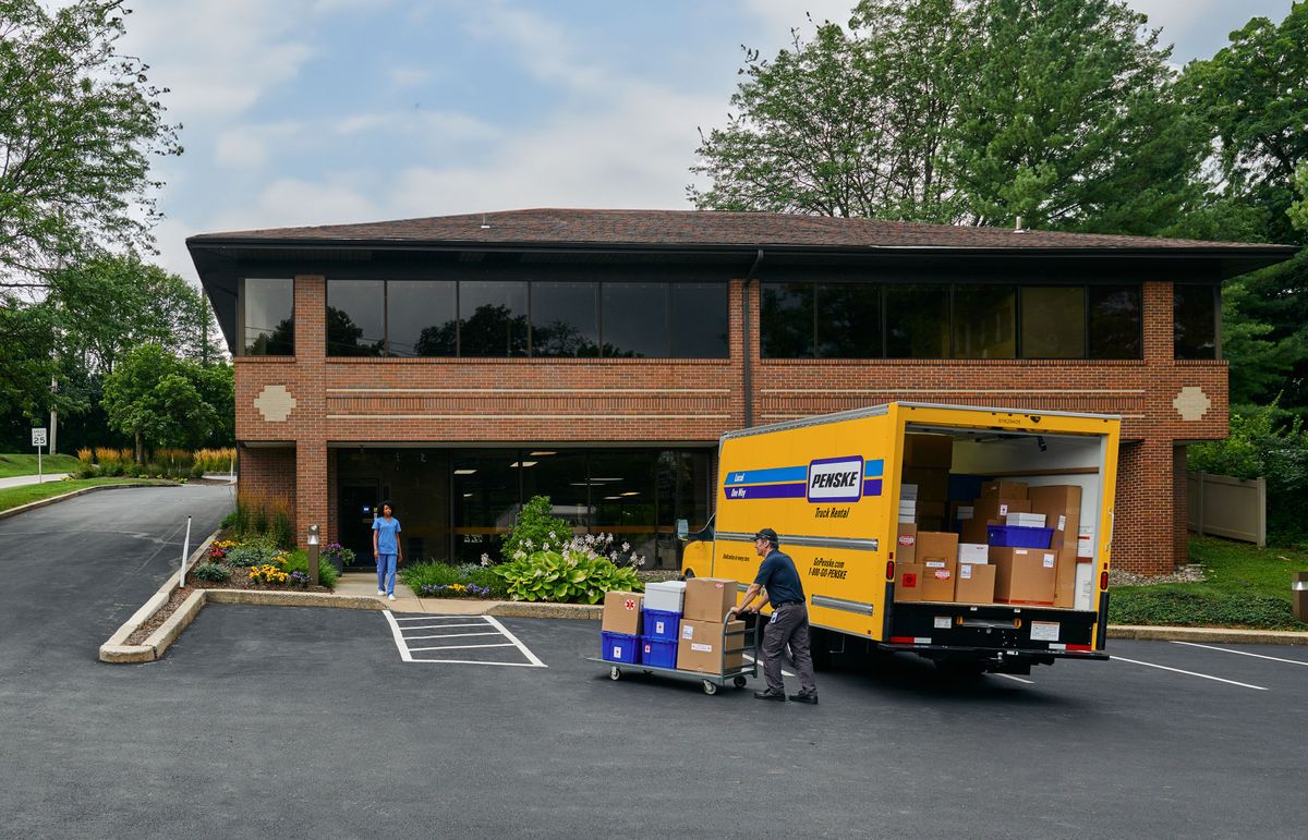 A delivery driver delivers boxes from a yellow Penske truck into a medical office. 