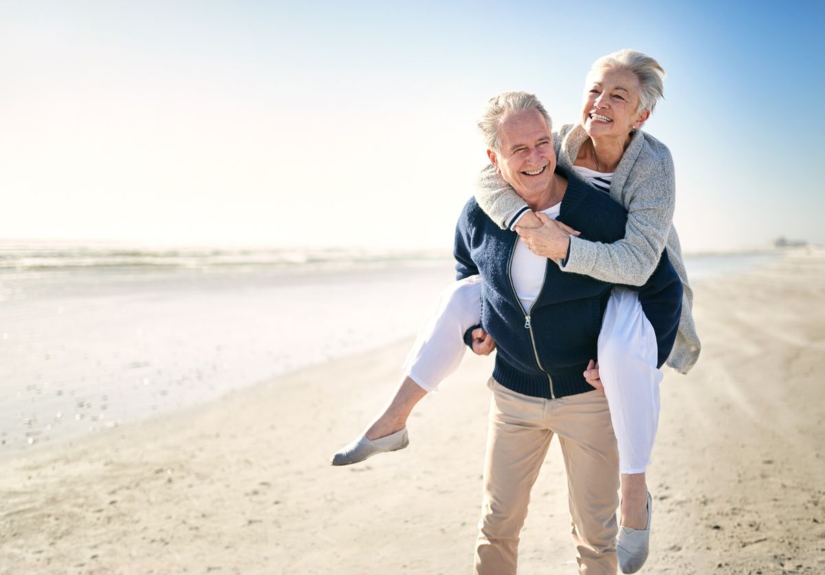 An older man carrying an older woman on his back, laughing on the beach.